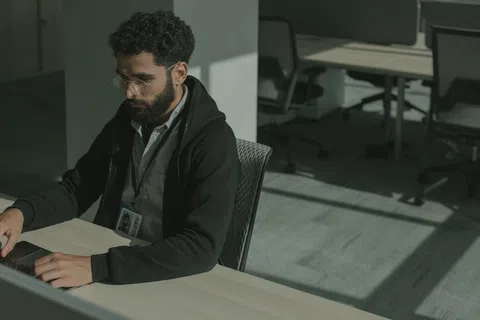 Man with curly hair and glasses working on a laptop in a modern, sunlit office space.