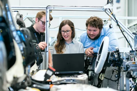 Three engineers collaborate on a laptop inside a vehicle assembly, focusing on automotive technology development.