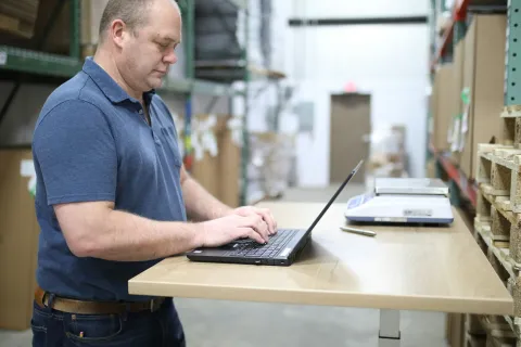 Man working on a laptop in a warehouse with shelves of pallets and packages around him.