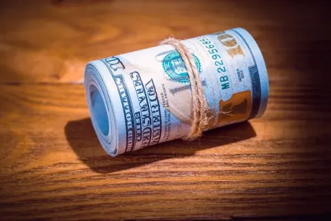 Rolled bundle of US 100 dollar bills tied with a string on a wooden surface.