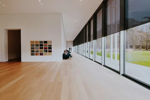 Modern art gallery with large windows, light wood floors, and visitors seated along white walls.