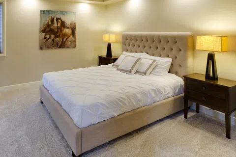 Cozy beige bedroom with tufted bed, white bedding, two nightstands, lamps, and horse painting on wall.