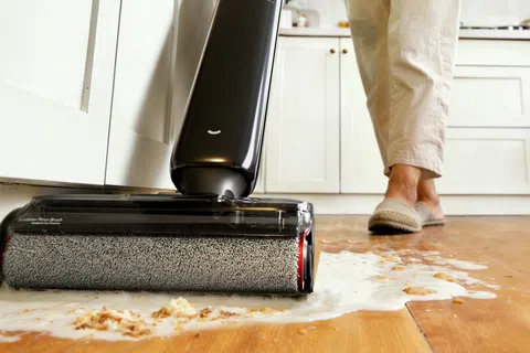 Person using an electric floor cleaner to mop spilled juice and crumbs off a hardwood kitchen floor.
