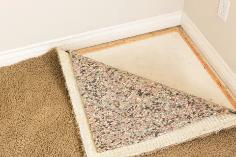Beige carpet being lifted to reveal floor padding and bare subfloor near a white baseboard and electrical outlet.
