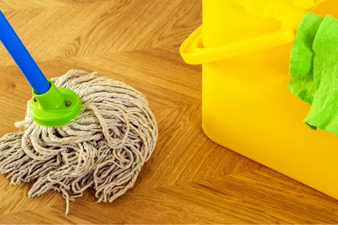 Wet mop head resting on wooden floor next to bright yellow bucket with green cleaning cloth.