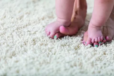 Close-up of baby feet standing on adult feet with green painted toenails on a soft cream rug.