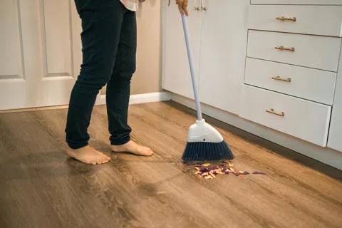 Person sweeping trash on wooden kitchen floor barefoot near white cabinets and drawers.