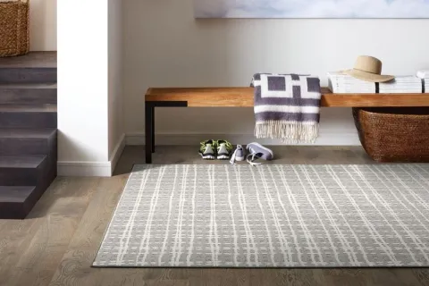Modern entryway with wooden bench, patterned rug, woven basket, folded blanket, shoes, and hat under soft natural light