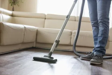 Person vacuuming a wooden floor next to a beige sectional sofa in a bright living room.
