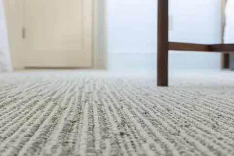 Close-up of textured beige carpet with wooden chair legs and white door in background inside bright room