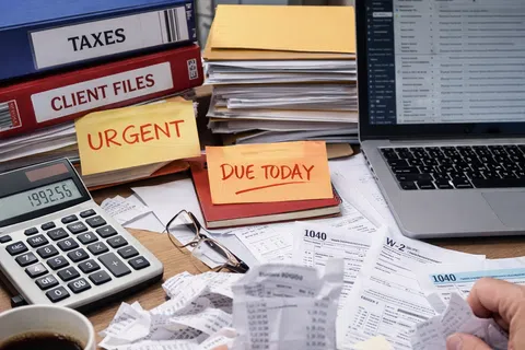 Desk cluttered with tax documents, calculator, laptop, urgent and due today notes, and client file folders.