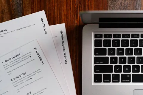 Close-up of a laptop keyboard beside printed user persona and brand color survey documents on a wooden desk