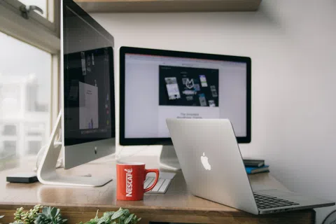 Workspace with Apple desktop and laptop, red Nescafe mug, and a wooden desk near window with plants.