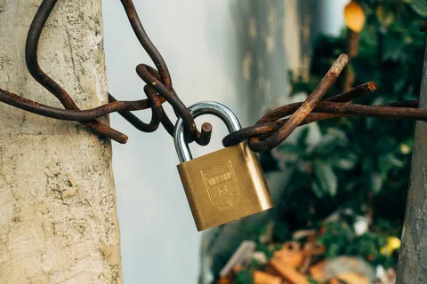 Brass padlock securing twisted rusty metal chains around a concrete post outdoors with greenery in background
