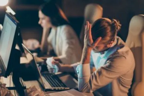 Man frustrated at work with head in hand and woman working on computer in background during late night.