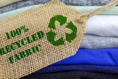 Stack of colorful folded clothes with a burlap tag labeled 100% recycled fabric and a recycling symbol.