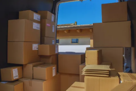 View of stacked cardboard boxes inside a delivery van with a building and blue sky outside the open door