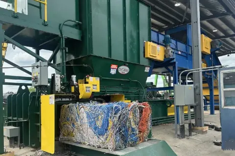 Industrial machine compressing a large colorful bale of mixed electrical wires inside a recycling facility.