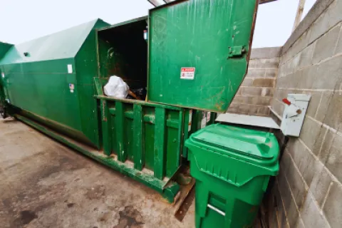 Large green commercial trash compactor with open lid and a nearby green waste bin in a concrete enclosure.