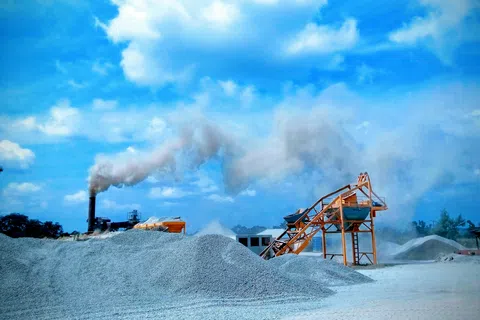 Construction site with gravel piles, conveyor belt, and factory smoke emitting under blue sky with clouds.