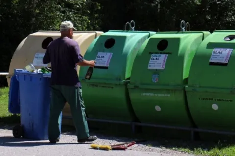 Man recycling glass bottles at green and beige recycling bins outdoors on a sunny day.