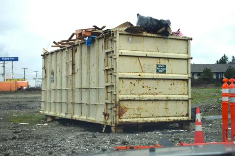 Large rusty dumpster labeled for wood recycling filled with scrap wood and debris on gravel lot.