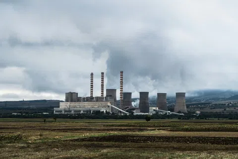 Industrial power plant with tall smokestacks emitting smoke under a cloudy sky near open fields.