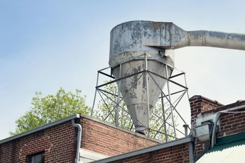 Rusty metal industrial silo with pipe on brick building roof against clear sky and green trees.