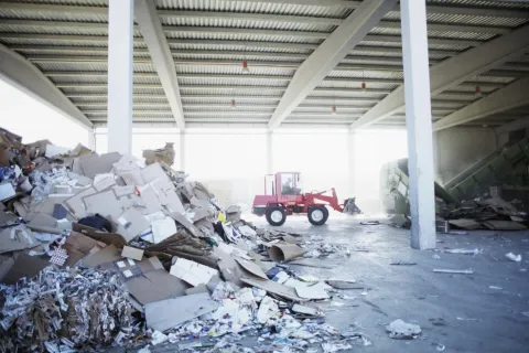 Indoor recycling facility with piles of cardboard and paper waste and a red loader vehicle clearing debris.