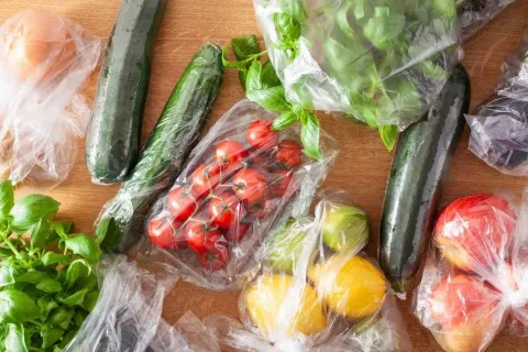 Fresh vegetables including zucchinis, cherry tomatoes, lemons, and basil wrapped in plastic bags on a wooden surface