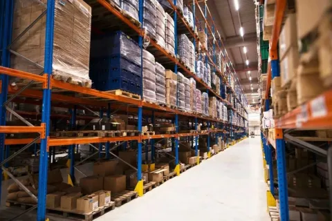 Large warehouse aisle with tall shelves stocked with pallets of boxes and goods under bright lighting