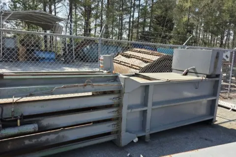 Large industrial metal baler machine placed outdoors near chain link fence and trees on a sunny day