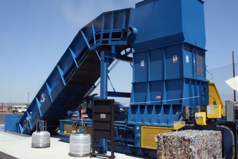 Large blue industrial machine compacting aluminum cans into bales outdoors on a sunny day