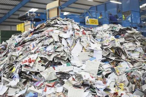 Large pile of mixed paper waste in a recycling facility with workers and machinery in the background