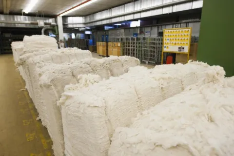 Large bales of raw cotton stacked in a spacious industrial warehouse with metal racks and yellow crates
