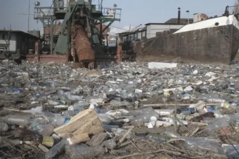 Polluted waterway filled with plastic waste near industrial ships highlighting environmental packaging challenges.