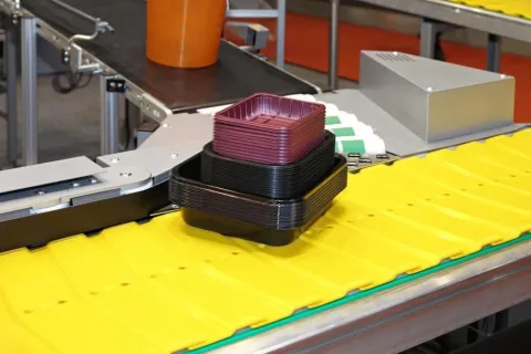Stacked black and purple plastic trays on a yellow conveyor belt in a manufacturing facility.