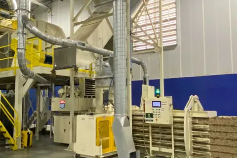 Industrial machinery in a factory setting with metal pipes, control panels, and stacked compressed materials.