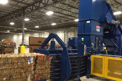 Industrial cardboard baler machine compressing recycled cardboard bales inside a warehouse facility.