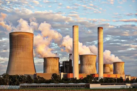 Large industrial power plant with cooling towers and chimneys emitting steam under a cloudy sky.