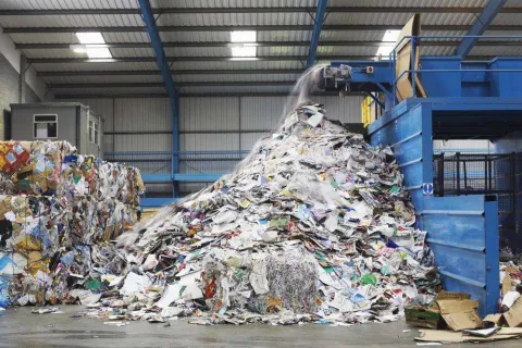 Large heap of mixed paper waste being processed by blue recycling machinery inside a warehouse facility