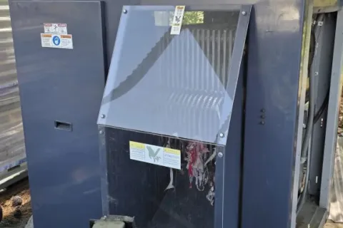 Large industrial gray machine with transparent front panel outdoors near trees under a metal roof.