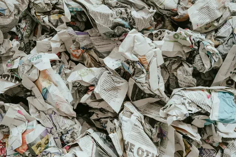 Pile of crumpled newspapers and printed paper waste in various colors and text scattered together.