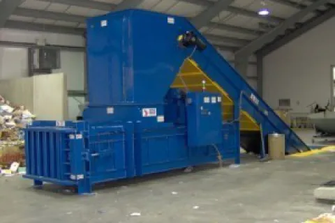 Large blue industrial waste baler machine inside a warehouse with conveyor belt for recycling materials.