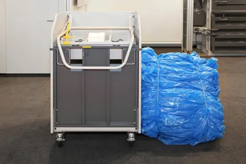 Gray industrial machine on wheels next to a large bundle of blue plastic sheets inside a factory.