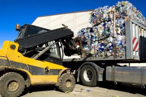 Yellow forklift loading compressed plastic bottle bales onto a truck for recycling under clear blue sky