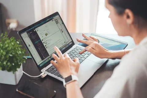 Person using laptop with messaging app open, typing on keyboard at a desk with plant and smartphone nearby