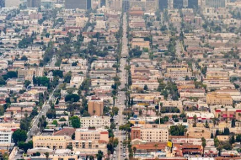 Aerial view of a sprawling urban cityscape with lined streets, various buildings, and distant skyscrapers under a hazy sky.