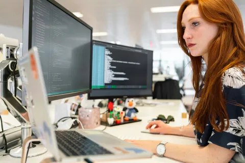 Focused woman with red hair coding on dual monitors at a bright office desk with laptop and tech accessories.