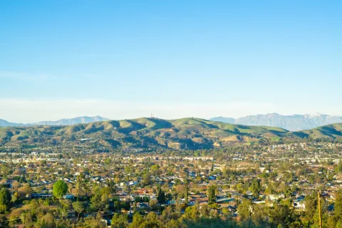 Panoramic view of a city with houses, trees, and green hills under a clear blue sky.